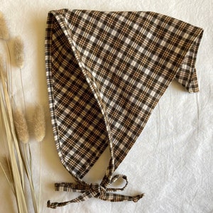 Puede incluir: Un pañuelo de bandana de cuadros marrones y blancos con un nudo atado en la parte inferior. El pañuelo está doblado en forma de triángulo.