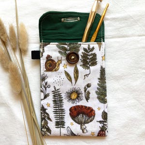 May include: A green fabric pouch with a white floral print featuring ferns, mushrooms, and snails. The pouch has a flap that closes with a button and is designed to hold knitting needles or crochet hooks.