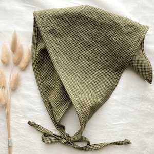 Puede incluir: Un pañuelo triangular de tela verde oliva con un patrón de cuadrícula blanca fina. El pañuelo cuenta con cintas de tela largas y delgadas en las esquinas inferiores para sujetarlo, con un dobladillo acabado a lo largo de los bordes.