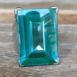 May include: A rectangular, teal gemstone ring with a silver setting. The gemstone has a faceted cut, reflecting light. The ring is set on a wooden surface.