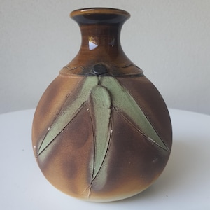 May include: A round ceramic vase with a glossy brown neck and a matte brown body. The vase features a raised, light green, three-leaf botanical design on the front. The surface has a smooth, glazed finish with subtle colour gradients throughout.