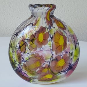 May include: A spherical glass vase featuring a clear base with swirling purple and green accents. The surface is decorated with multiple floral motifs in shades of muted pink and yellow, integrated into the glass structure with a smooth finish.
