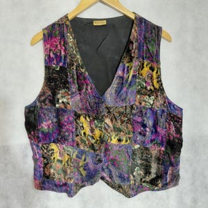 Puede incluir: Un chaleco sin mangas con un diseño de patchwork de tela de terciopelo en tonos morado, negro, verde y amarillo. La prenda tiene un escote en V, un forro interior negro y tres botones oscuros en la parte delantera. Una pequeña etiqueta es visible.