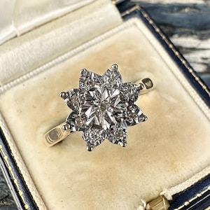 Puede incluir: Un anillo de oro vintage con un racimo floral de diamantes. El anillo, engastado en una banda de oro, se presenta en un joyero azul y crema. Los diamantes brillan.