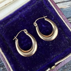 Könnte beinhalten: Ein Paar goldene Creolen mit glatter, polierter Oberfläche und abgerundetem U-förmigem Design. Die Ohrringe verfügen über einen Klappverschluss und werden in einer Schmuckschatulle mit dunkelviolettem Samtfutter präsentiert.