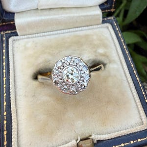 Puede incluir: Un anillo tipo cluster con un diamante central de talla brillante rodeado por un halo de diamantes redondos más pequeños en una banda de metal plateado. El anillo se presenta dentro de un joyero cuadrado azul oscuro con forro de terciopelo crema.