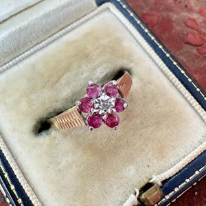 May include: A floral ring featuring a central diamond and six surrounding ruby gemstones. The ring has a gold band with a textured pattern. Presented in a blue jewellery box with a cream interior.