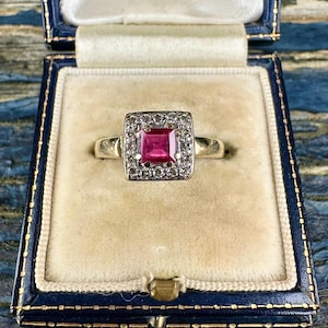 Puede incluir: Un anillo de oro con una gema rosa brillante cuadrada en el centro, rodeada por un halo de piedras más pequeñas y claras. El anillo se exhibe en un joyero azul con interior color crema.