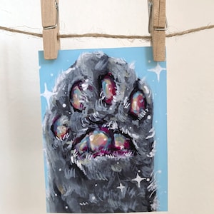 Op de afbeelding: Een close-up van een grijze kattenpoot met roze en blauwe voetkussentjes. De poot is geschilderd op een lichtblauwe achtergrond met witte glitteraccenten.