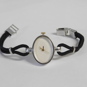 May include: An oval-faced wristwatch with a silver-tone case and gold-tone crown. The watch has a white face with gold hands and is attached to a black rope-style band with silver clasps. The watch is displayed on a white surface.