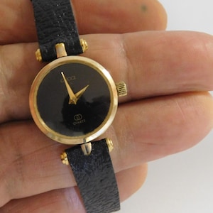 May include: A wristwatch featuring a round gold-tone case and a black dial with gold-tone hands. The watch has a textured black strap and a small gold-tone crown on the side. The dial displays the brand name Gucci and the word Quartz.