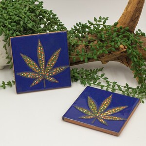 Könnte beinhalten: Zwei blaue Keramik-Untersetzer mit einem goldenen und orangefarbenen Marihuana-Blatt-Design. Die Untersetzer befinden sich auf einer weißen Oberfläche.