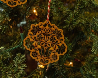 May include: A wooden Christmas ornament with a floral design, featuring a tree with birds and flowers. The ornament is painted in a warm brown color and is hanging on a Christmas tree.