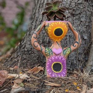 May include: A terracotta planter shaped like a human figure with painted flowers and a heart. The planter is holding a succulent plant in its head.