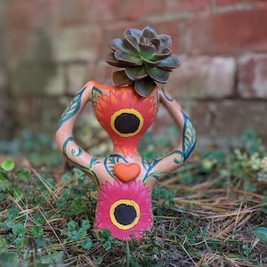 May include: A terracotta planter shaped like a human figure with painted details. The planter has two large orange flowers with black centres and a smaller pink flower with a black centre. A small succulent plant is growing out of the top of the planter.