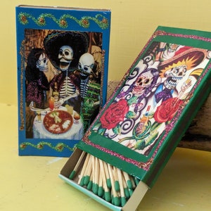 May include: Two matchboxes with colorful designs featuring skeletons and flowers. The top box is blue with a green border and the bottom box is green with a pink border. The bottom box is open and shows the matches inside.