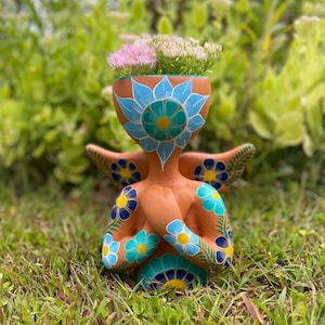 Könnte beinhalten: Ein terrakottafarbener Keramik-Blumentopf in Form einer Figur, verziert mit blauen und türkisfarbenen Blumenmustern. Der Pflanztopf enthält rosa und grüne Blütenpflanzen. Die Figur hat ausgestreckte Arme und sitzt im Gras.