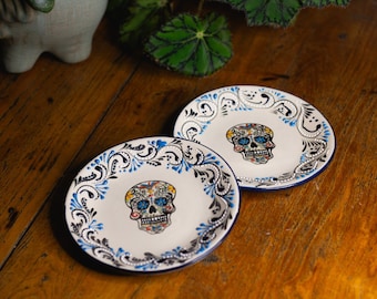 May include: Two white ceramic plates with blue and black floral designs around the edges. Each plate features a colorful sugar skull design in the center.