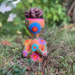 May include: A terracotta-colored planter shaped like a human figure with hands clasped at the chest. The planter is decorated with hand-painted blue, purple, and orange flowers and green vines. A dark purple succulent is planted in the top opening.