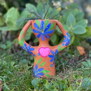 May include: A terracotta-colored ceramic planter in the shape of a person, with arms raised. It features blue flower and green leaf designs, a pink heart, and holds a green plant. The planter is set in a grassy outdoor environment.