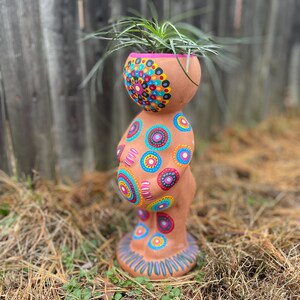 May include: A terracotta-colored ceramic planter in the shape of a stylized figure. The planter is adorned with colorful, hand-painted circular patterns in shades of blue, pink, yellow, and green. The planter holds a green plant with long, thin leaves.