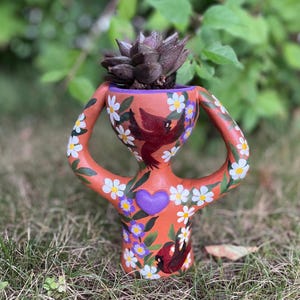 May include: A terracotta-coloured anthropomorphic planter featuring two curved arms, painted with white daisies, purple flowers, and red birds. A purple heart is centred on the torso, and a dark succulent is planted in the top opening.