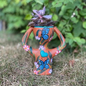 May include: A terracotta-colored anthropomorphic planter with two curved handles, painted with blue butterflies, pink and white flowers, and a central turquoise heart. A purple succulent plant is potted inside the top opening of the vessel.