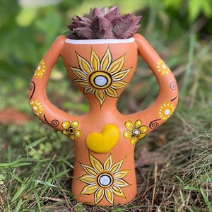 May include: A terracotta-colored ceramic planter shaped like a stylized figure with raised arms. The surface features hand-painted yellow sunflowers, black swirling accents, and a raised yellow heart on the chest. A dark purple succulent sits inside.