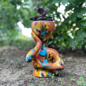 May include: A terracotta-colored ceramic planter shaped like a human bust with one hand touching the face. The surface is hand-painted with yellow and white flowers, green leaves, and blue blossoms. A bright blue heart is attached to the chest.