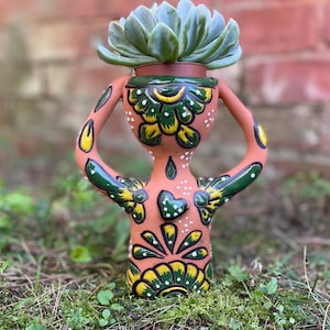 May include: A terracotta-colored ceramic planter in the shape of a person, holding its arms up. The planter is decorated with green and yellow floral designs and white dots. A succulent plant sits in the top of the planter.