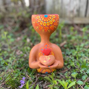Könnte beinhalten: Eine terrakottafarbene Keramikfigur mit großem Kopf und einem Körper, der ein herzförmiges Objekt hält. Kopf und Brust sind mit farbenfrohen Mustern im Mandala-Stil in Gelb, Orange und Blau verziert. Die Figur befindet sich im Freien, umgeben von grünem Gras und Laub.