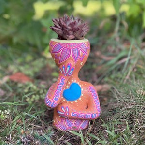 May include: A terracotta-colored planter shaped like a human torso with a succulent planted in the top. The planter features intricate hand-painted floral and geometric patterns in pink, blue, and white, with a raised blue heart on the chest.