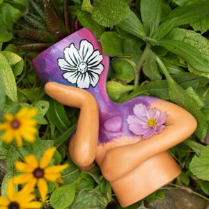 Könnte beinhalten: Ein keramischer Pflanztopf in Form einer Figur, bemalt in Lila- und Beigetönen. Ein weißes Blumendesign ziert den Kopf, und eine rosa Blume ruht auf der Brust. Der Pflanztopf ist von grünem Laub und gelben Blumen umgeben.