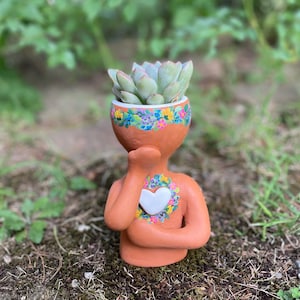 Puede incluir: Una maceta de cerámica de color terracota con forma de persona, sosteniendo una mano en la barbilla. La maceta presenta un diseño de corazón blanco y un borde floral alrededor de la parte superior, con una planta suculenta.