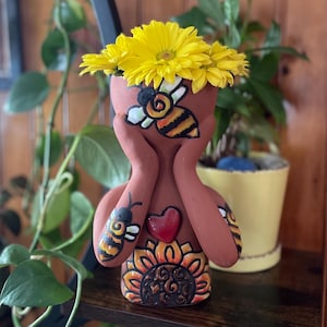 Puede incluir: Una maceta de cerámica de color terracota con forma de figura, adornada con un diseño pintado de abeja y girasol. La maceta contiene flores de color amarillo brillante. Un corazón rojo está pintado en el pecho de la figura.