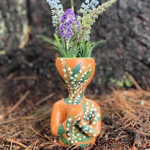 May include: A terracotta-colored ceramic vase in the shape of a person, decorated with white and green floral patterns. The vase holds a bouquet of purple and blue artificial flowers. The vase is sitting on a bed of pine needles.