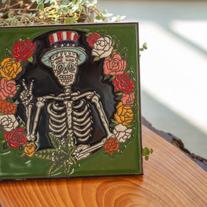 May include: A decorative square tile featuring a skeleton wearing a top hat with red, white, and blue stripes, surrounded by a wreath of colorful roses. The skeleton is giving a peace sign and holding a cannabis leaf. The tile has a green border.