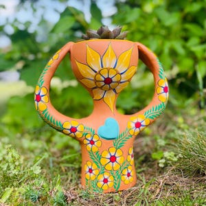 May include: A terracotta-colored ceramic planter shaped like a stylized figure with two arched handles. The surface is hand-painted with yellow flowers featuring red centers, green leafy vines, and a single raised light blue heart on the chest.