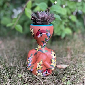 May include: A terracotta-colored anthropomorphic planter shaped like a human torso and head, featuring hand-painted yellow flowers, purple blossoms, and red ladybugs. The rim is painted turquoise, and a dark purple succulent sits inside the top.