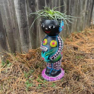 May include: A black, hand-painted ceramic planter in the shape of a stylized figure. The planter features colorful floral designs in pink, yellow, and blue. The figure holds a plant with long green leaves. The planter is standing on a base of dry grass.