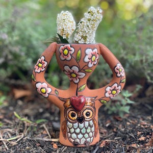 May include: A terracotta-colored ceramic vase shaped like a stylized figure with two arched arms. The surface features hand-painted white flowers with yellow centers and a central owl illustration with a red heart above its head, holding white flowers.