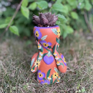 May include: A terracotta-colored planter shaped like a figure with hands held to its face, featuring hand-painted purple pansies, yellow daisies, green leaves, and a central purple heart. A dark-colored succulent is planted in the top opening.