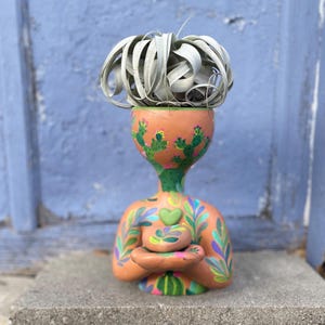 Könnte beinhalten: Ein terrakottafarbener Keramik-Blumentopf in Form einer Person, verziert mit bemalten Kakteen und Blättern. Der Pflanztopf enthält eine graue Luftpflanze. Die Figur hat die Arme verschränkt und hält eine kleine Schale. Das Design umfasst grüne, blaue und rosa Akzente.