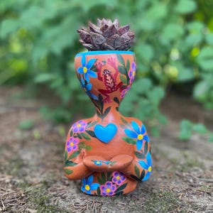 May include: A terracotta-colored planter shaped like a human bust, featuring hand-painted blue flowers, pink blossoms, green leaves, a red frog, and a central blue heart. A dark, spiky succulent plant sits inside the top opening of the planter.