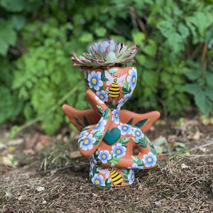 May include: A terracotta-colored ceramic planter shaped like a figure, adorned with blue floral and bee designs. The planter holds a succulent plant with green and purple leaves. The figure has outstretched arms and a heart-shaped detail.