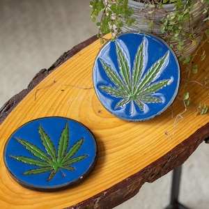 Könnte beinhalten: Zwei blaue Keramik-Untersetzer mit einem grünen Marihuana-Blatt-Design. Die Untersetzer befinden sich auf einer Holzoberfläche.