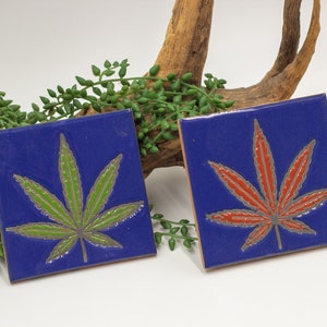 May include: Two square ceramic tiles with a blue background and a green and red marijuana leaf design.