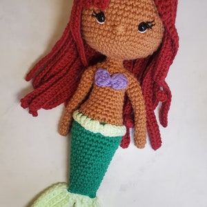 May include: A crocheted mermaid doll with red hair, a brown face, a purple seashell bra, a green tail, and a light green tail fin.