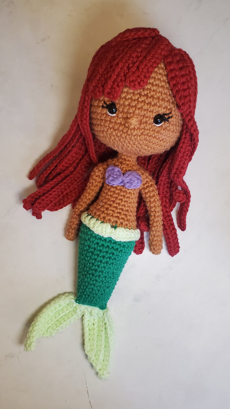 May include: A crocheted mermaid doll with red hair, a brown face, a purple seashell bra, a green tail, and a light green tail fin.
