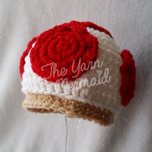May include: A crocheted mushroom hat with a red top, white body, and brown brim. The hat is made with yarn and has a textured surface. The text "The Yarn Mermaid" is visible on the hat.